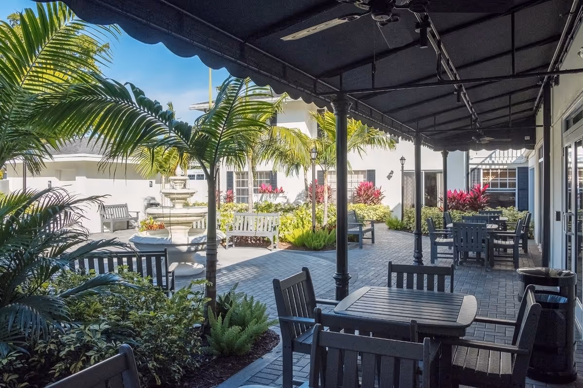 Outdoor patio area with black wooden tables and chairs under a black canopy. Surrounding the patio are lush green plants, palm trees, and colorful shrubs. In the background, there is a white building with windows and benches, and a multi-tiered stone fountain is visible in the garden area.