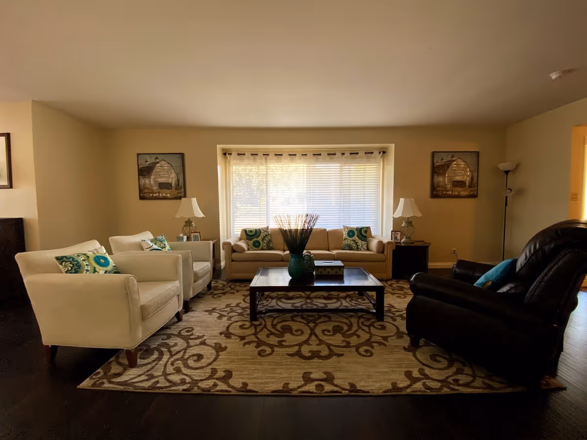 A cozy living room with a beige sofa and two matching armchairs with patterned cushions, a dark leather recliner, a wooden coffee table with decorative items, two side tables with lamps, framed pictures on the walls, and a large window with blinds and sheer curtains letting in natural light.