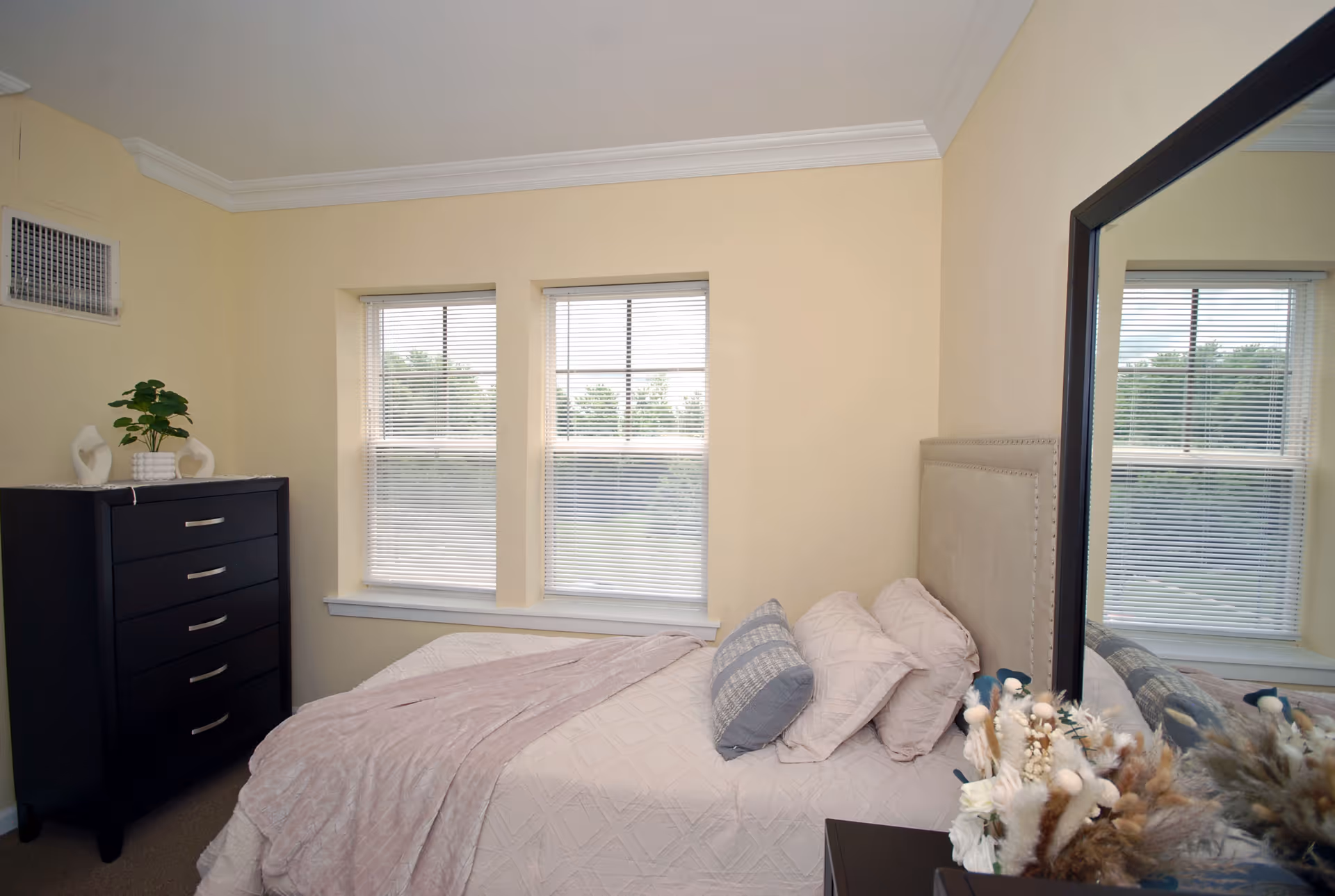 A small bedroom with a made bed, decorative pillows, a dresser, double windows with blinds, and a large mirror.