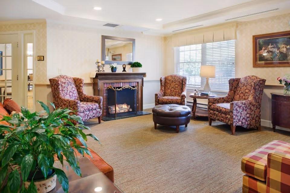 A cozy living room with a lit fireplace, three patterned armchairs, a round leather ottoman, a side table with a lamp, a large window with blinds, a framed painting on the wall, and a green potted plant in the foreground.