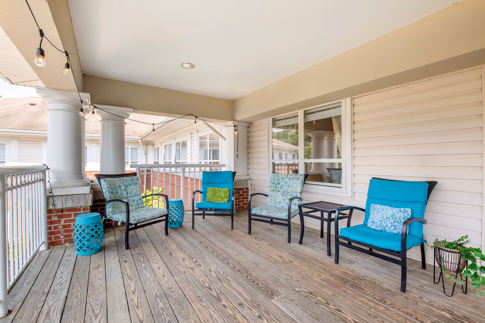 Covered outdoor patio area with wooden flooring, four cushioned chairs with blue and patterned cushions, two small blue side tables, string lights hanging from the ceiling, white railing, and beige siding walls with a window.