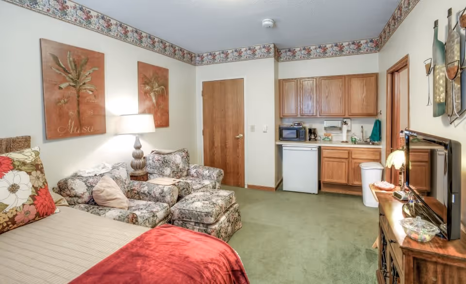 A cozy room featuring a bed with floral and red pillows, a floral patterned armchair with an ottoman, a wooden side table with a lamp, and two framed palm tree artworks on the wall. There is a small kitchenette area with wooden cabinets, a mini fridge, microwave, coffee maker, and sink. A wooden TV stand with a flat-screen TV and decorative items is on the right side. The room has green carpet and floral wallpaper border near the ceiling.