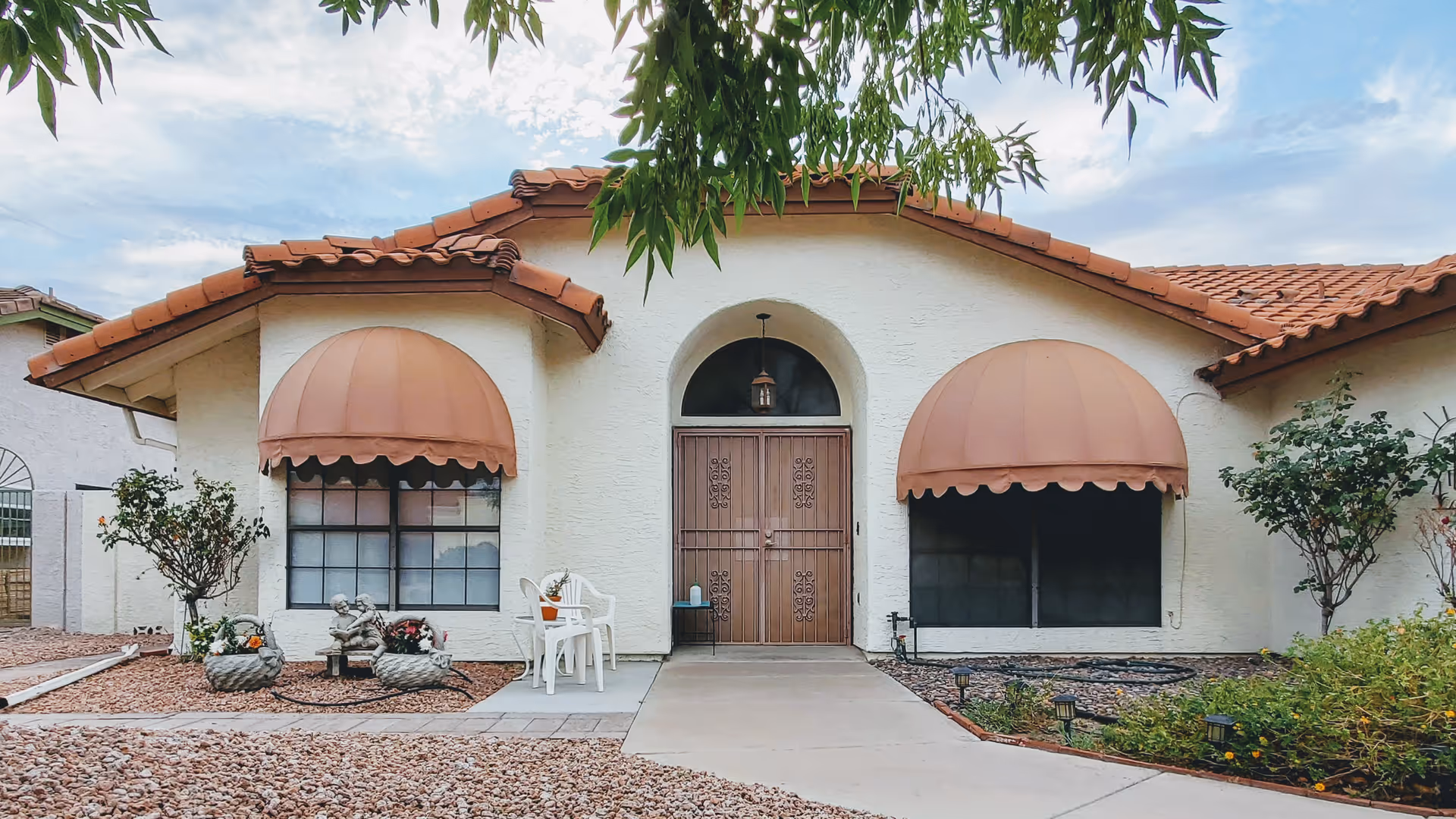 Front exterior of a single-story stucco house with terracotta tile roof, two rounded awnings over windows, a central double door, walkway, and small landscaped plants and chairs.