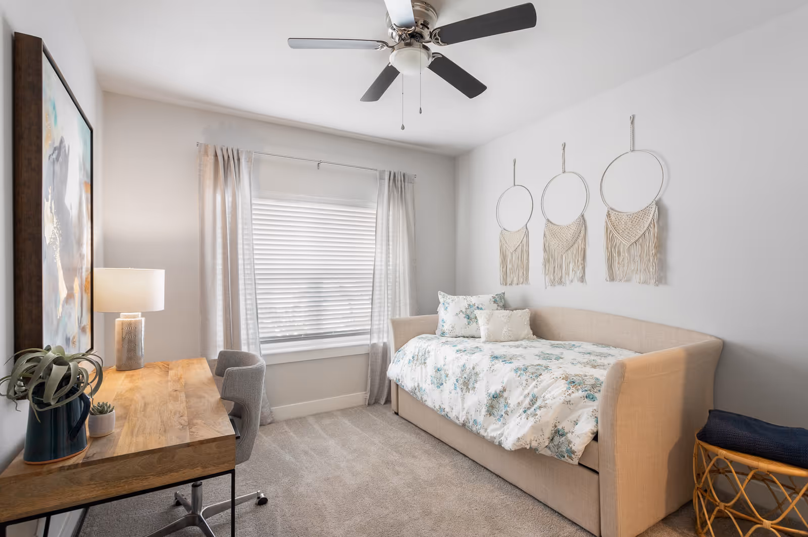A bright bedroom with a beige daybed covered in floral bedding and pillows, three circular macrame wall hangings above the bed, a wooden desk with a lamp and plants, a gray office chair, a window with white blinds and light curtains, and a ceiling fan with dark blades.