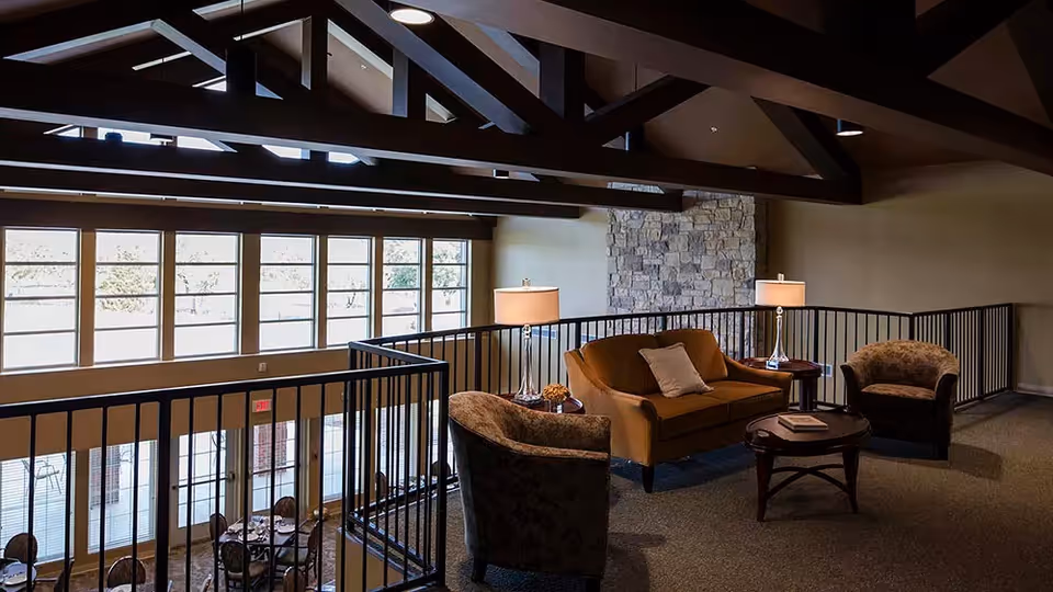 A cozy seating area in an assisted living facility with a brown sofa, two armchairs, a coffee table, and two table lamps. The space features large windows letting in natural light, exposed wooden ceiling beams, and a stone accent wall.