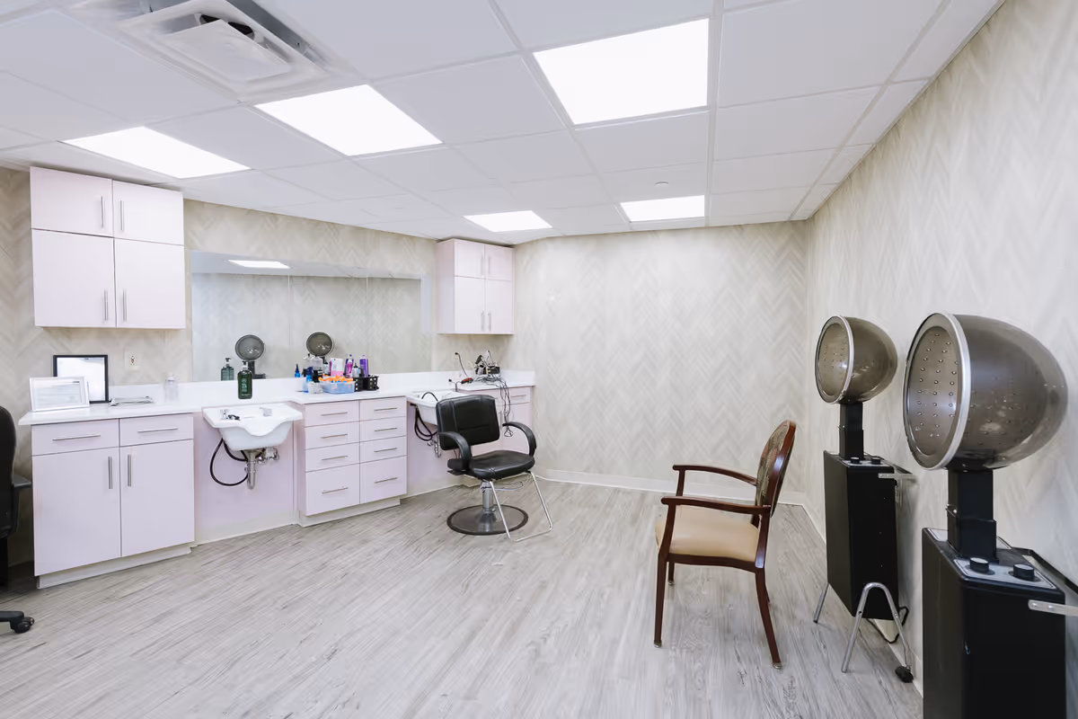 A clean and modern salon room with light wood flooring and chevron-patterned wallpaper. The room features a large mirror above a counter with cabinets and drawers, a small sink, a black salon chair, two vintage hair dryers on stands, and a wooden chair with a cushioned seat. The ceiling has recessed lighting panels.