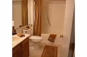A bathroom with a white toilet, a bathtub with a shower curtain, a beige bath mat on the floor, a wooden vanity with a sink, and a mirror above the sink. Towels are placed on the toilet tank and bathtub edge.
