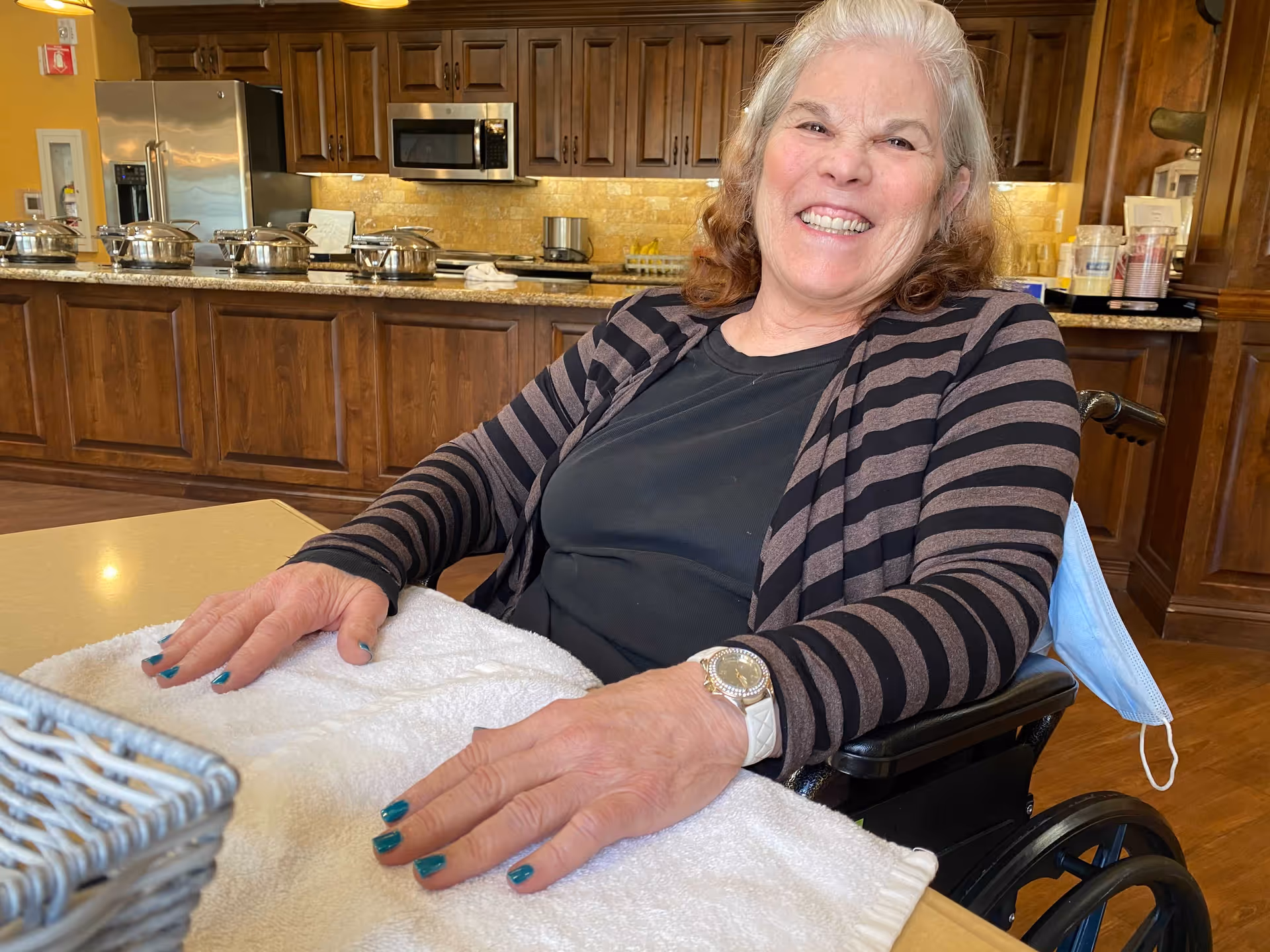 A smiling elderly woman with gray hair sitting in a wheelchair at a table covered with a white towel, in a kitchen area with wooden cabinets, stainless steel appliances, and several covered serving dishes on the counter behind her.