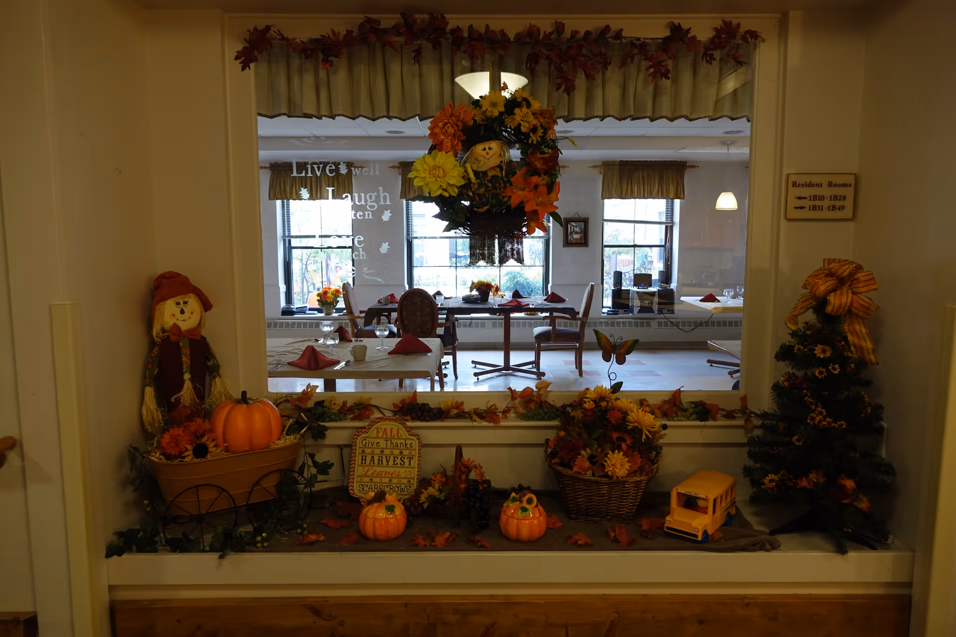 A window decorated with autumn-themed items including pumpkins, flowers, a scarecrow figure, and a small decorative tree with a bow. Through the window, a dining room is visible with tables set with red napkins and chairs, and large windows letting in natural light.