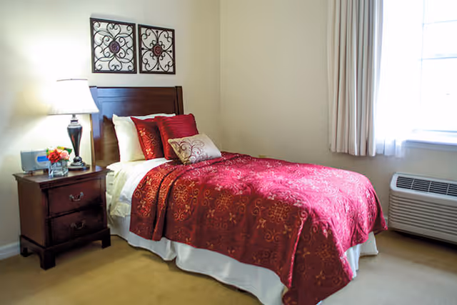 A bedroom with a single bed covered in a red and gold patterned bedspread and multiple pillows. There is a wooden nightstand with a lamp, a clock, and a small flower arrangement. Two decorative wall hangings are above the bed. A window with light curtains and an air conditioning unit is on the right side.
