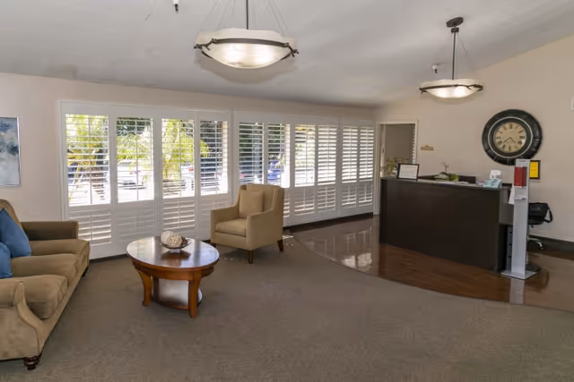 A reception area with a dark wooden front desk, a large wall clock, and a hand sanitizer dispenser. The room has beige walls, carpeted floor with a wooden section near the desk, a beige sofa with blue cushions, a beige armchair, and a round wooden coffee table. Large windows with white plantation shutters let in natural light.