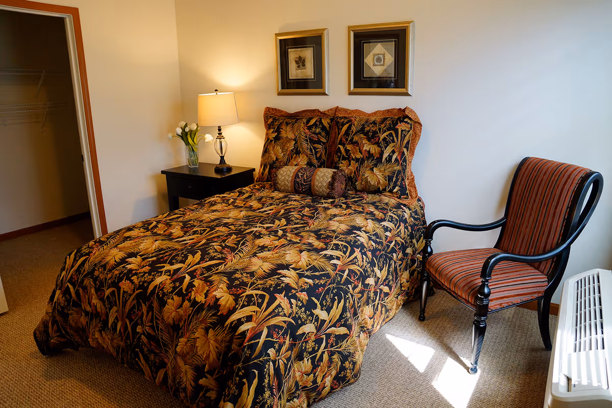 A bedroom with a bed covered in a dark floral patterned comforter and matching pillows. Next to the bed is a black nightstand with a lamp and a vase of white flowers. On the wall above the bed are two framed pictures. To the right of the bed is a striped upholstered armchair and a white air conditioning unit below the window. An open closet door is visible on the left side of the room.
