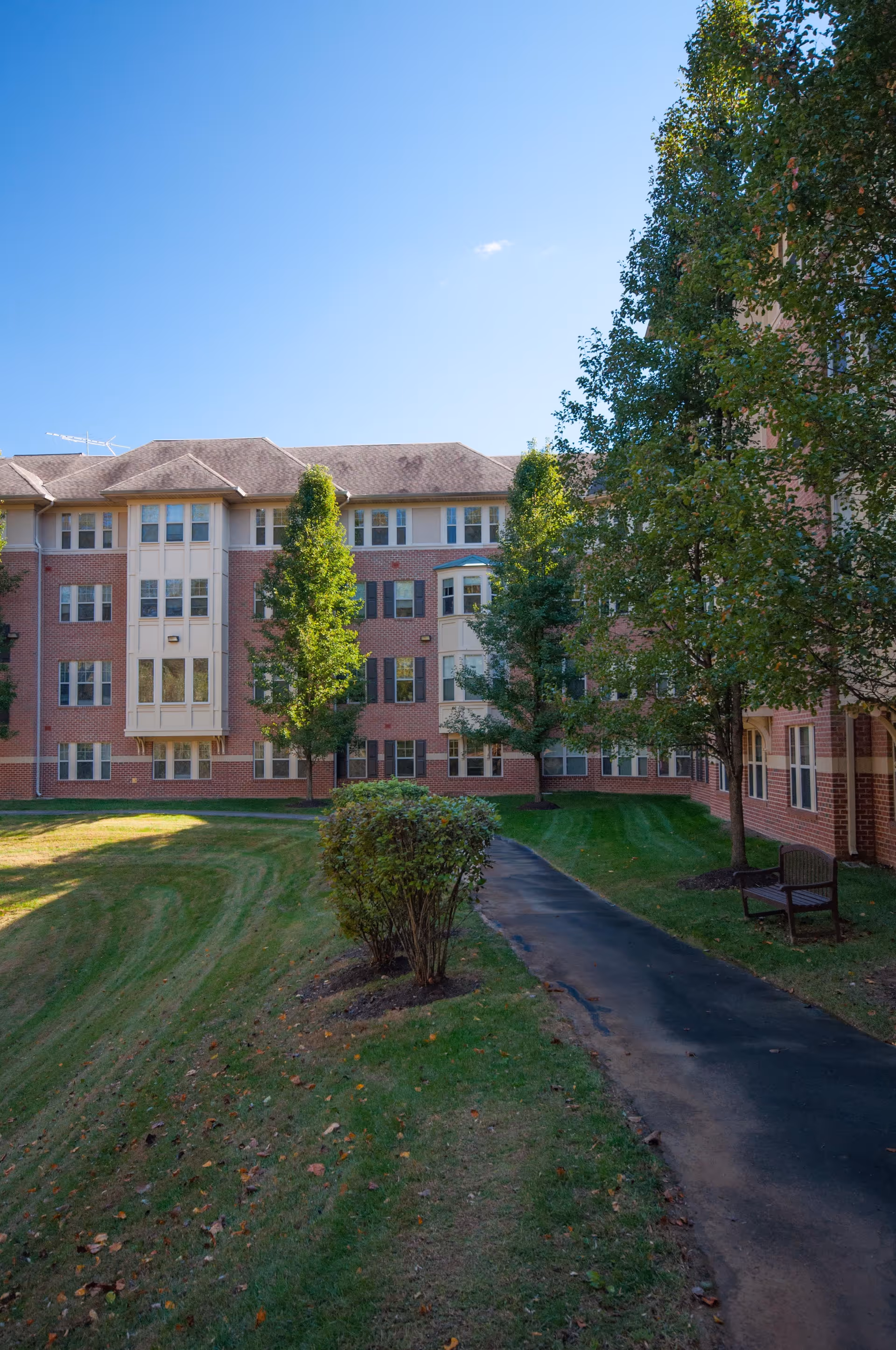 Exterior view of a multi-story brick senior living facility building with multiple windows, surrounded by green grass, trees, a paved walkway, and a bench under a clear blue sky.