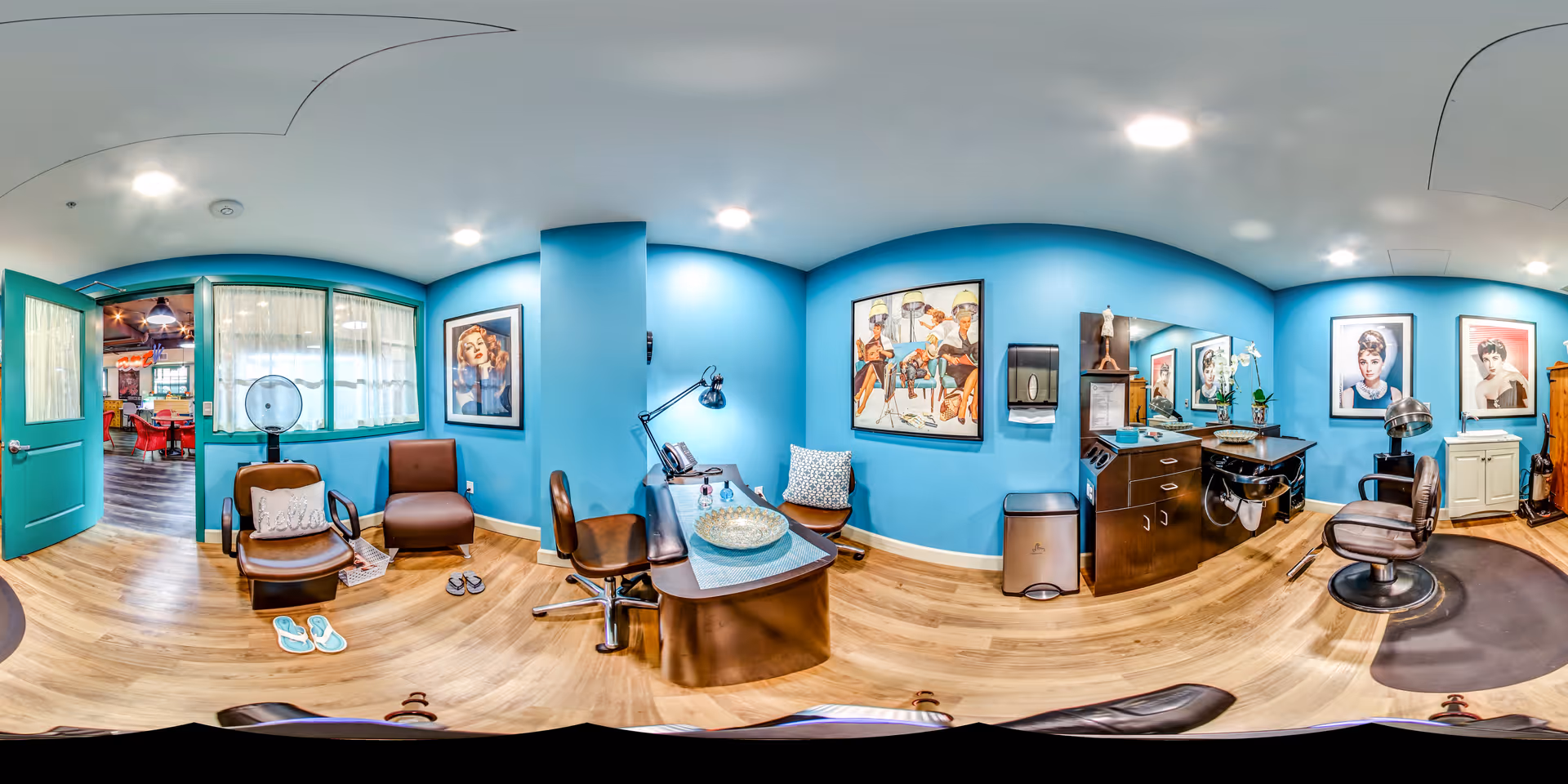 Interior of a salon room with turquoise walls and wooden flooring. The room features salon chairs, a manicure table with a lamp, a hair washing station, and framed vintage-style portraits on the walls. There are slippers on the floor and a door leading to another area with red chairs visible.