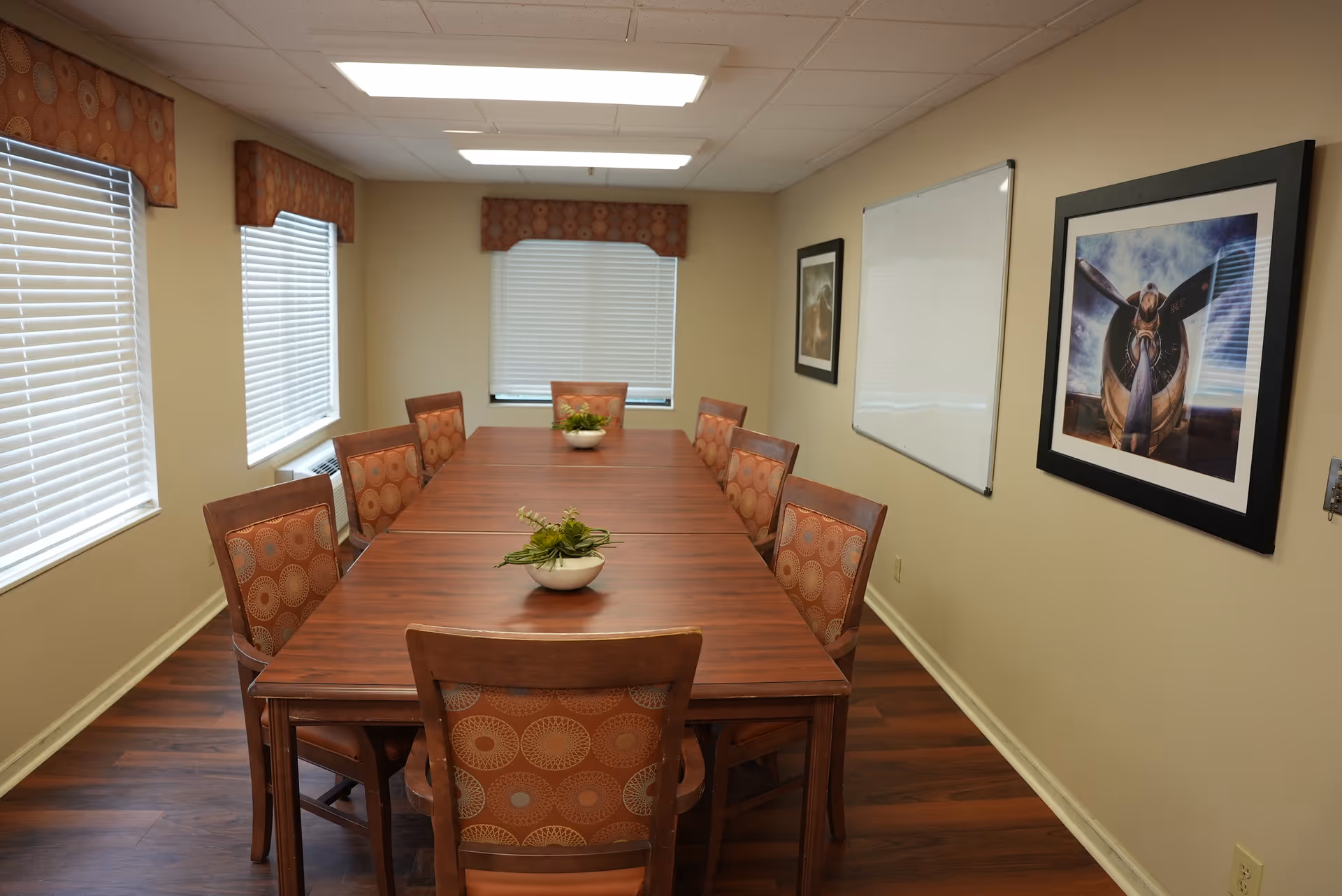 A long wooden dining/conference table with chairs in a bright room with windows, a whiteboard, and framed artwork on the wall.