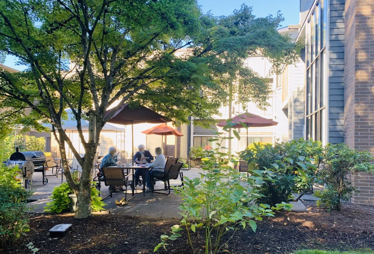 Outdoor courtyard area at Creekside Village Retirement Residence with several elderly people sitting around a table under large umbrellas. The area is surrounded by trees, bushes, and a building with large windows.