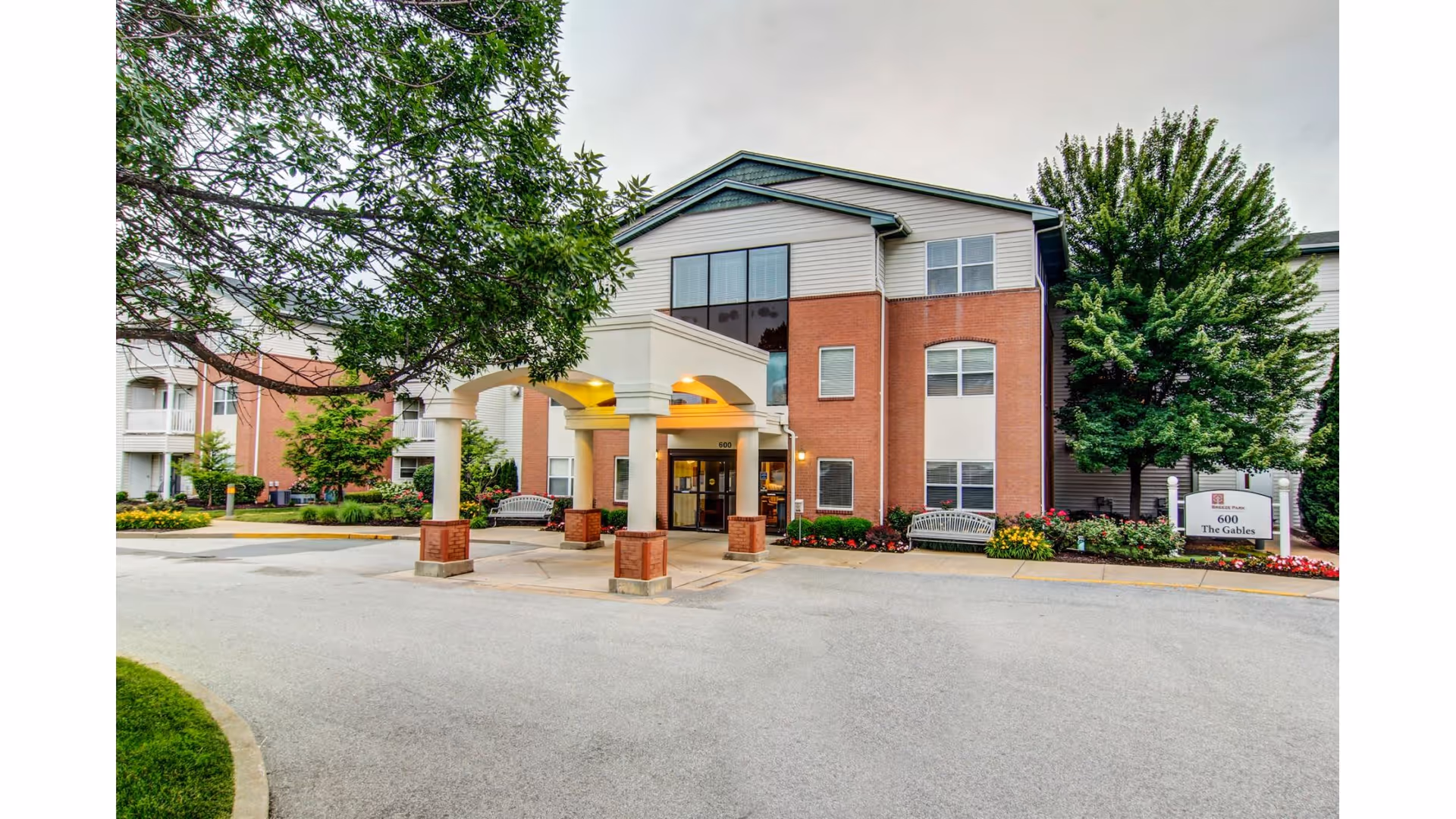 Exterior view of a three-story senior living facility building with a covered entrance supported by columns. The building has a combination of brick and siding exterior with large windows. There are benches and landscaped flower beds near the entrance, and trees surrounding the area. A sign near the entrance reads '600 The Gables'.