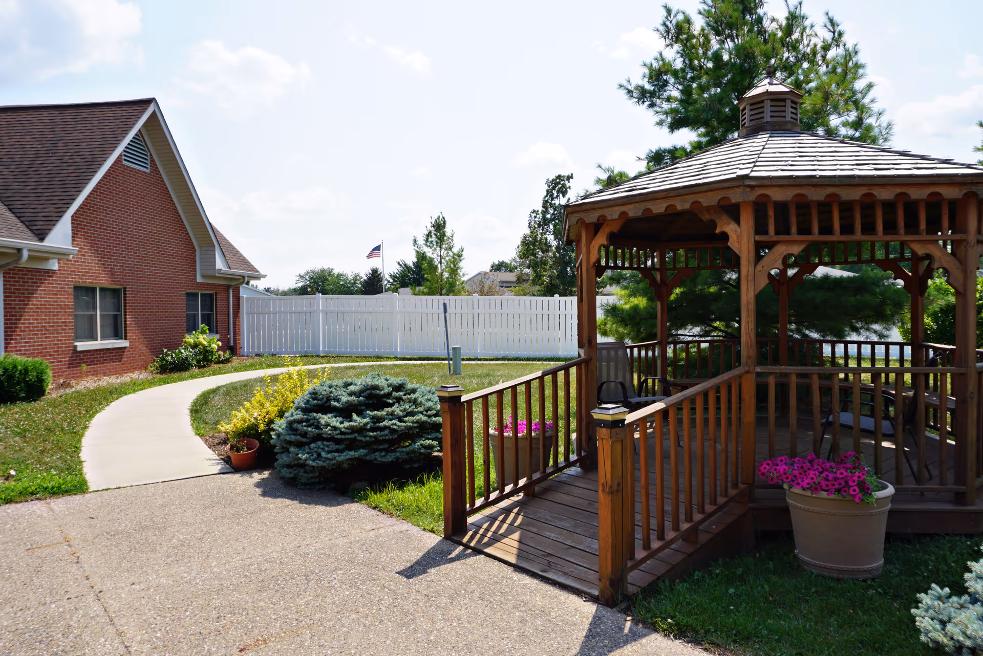 Outdoor view of a senior living facility showing a wooden gazebo with a ramp, surrounded by green grass, potted flowers, and shrubs. A brick building with windows and a white fence are visible in the background under a partly cloudy sky.