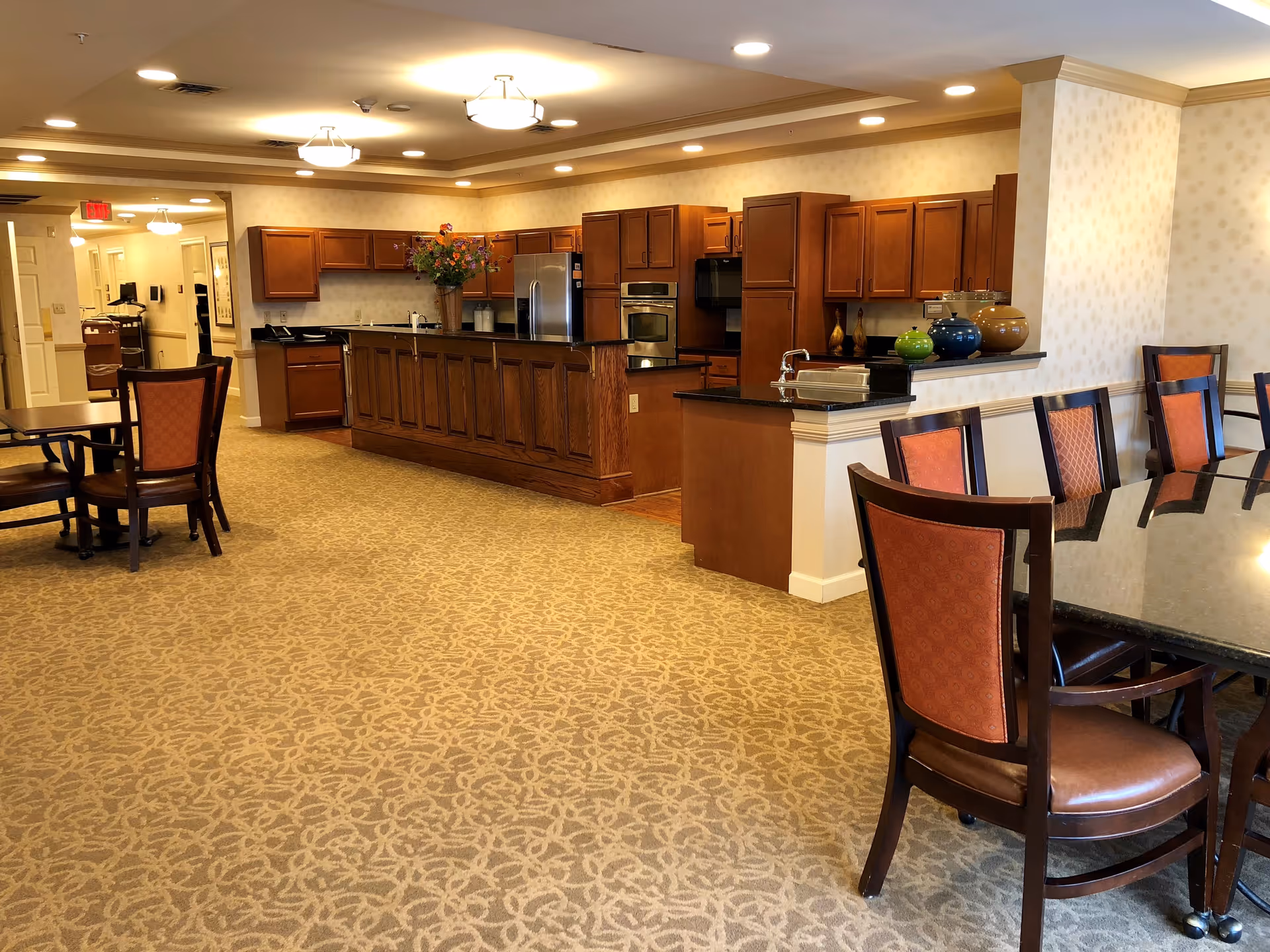 Interior view of a senior living facility dining area and kitchen. The room features a long kitchen counter with wooden cabinets, stainless steel appliances, and a sink. There are multiple dining tables with chairs that have orange upholstery. The carpet has a beige pattern, and the ceiling has recessed lighting and hanging light fixtures. A vase with flowers is placed on the kitchen counter.