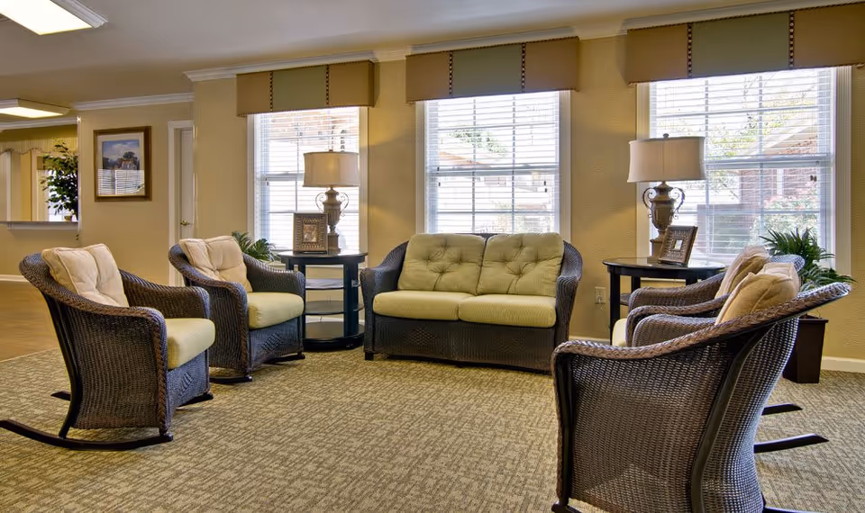 A cozy senior living facility common area with wicker furniture including a loveseat and four armchairs arranged in a circle on a carpeted floor. Two side tables with lamps and picture frames are placed between the windows, which have blinds partially open to let in natural light. The walls are painted beige and there is a framed picture hanging on the wall.