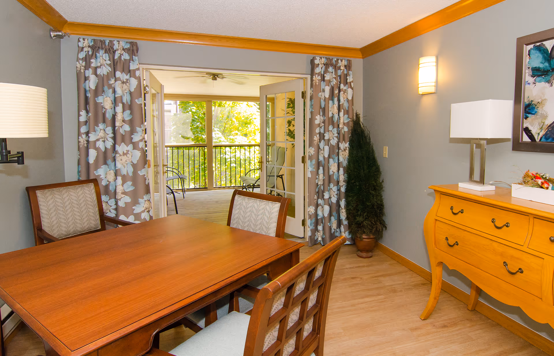 A dining area with a wooden table and four chairs, floral curtains framing glass doors that open to a balcony with outdoor chairs and greenery visible outside. The room has light gray walls, a wooden sideboard with a lamp and decorative items, and a potted plant in the corner.