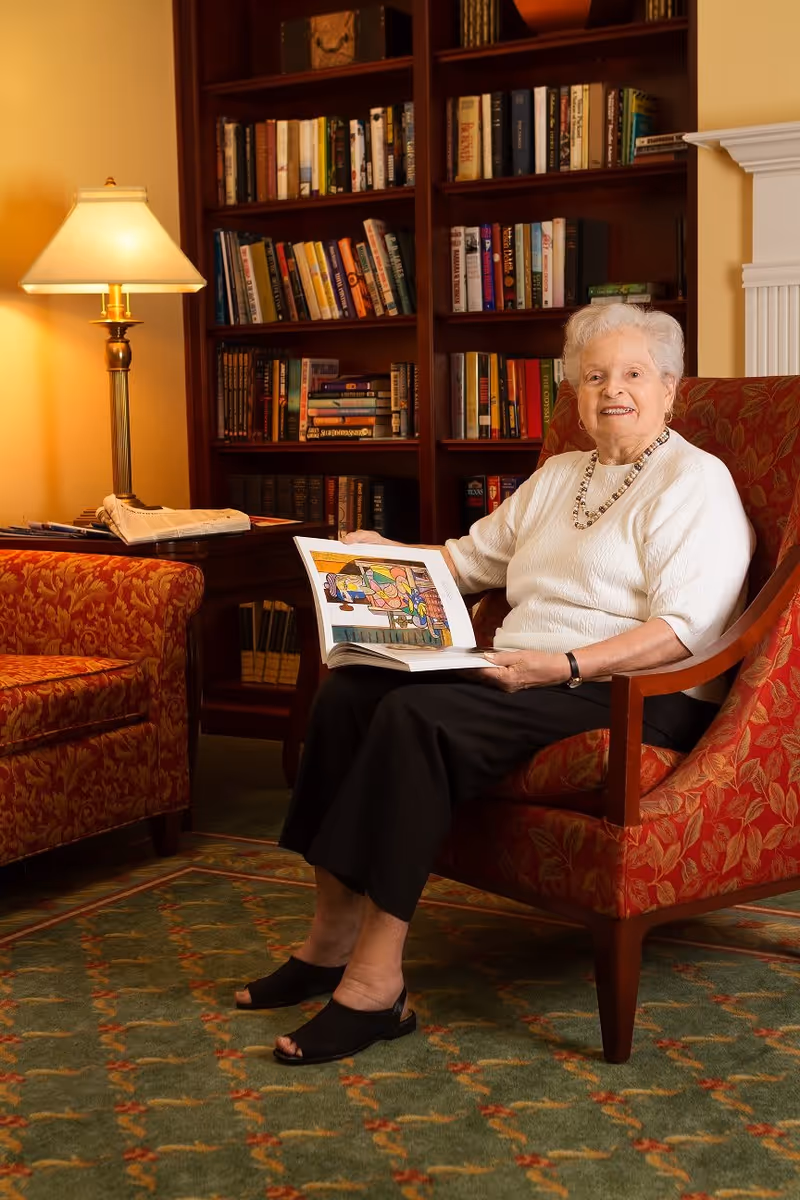 An elderly woman sits in a patterned armchair reading an open book in a cozy library-style living room.