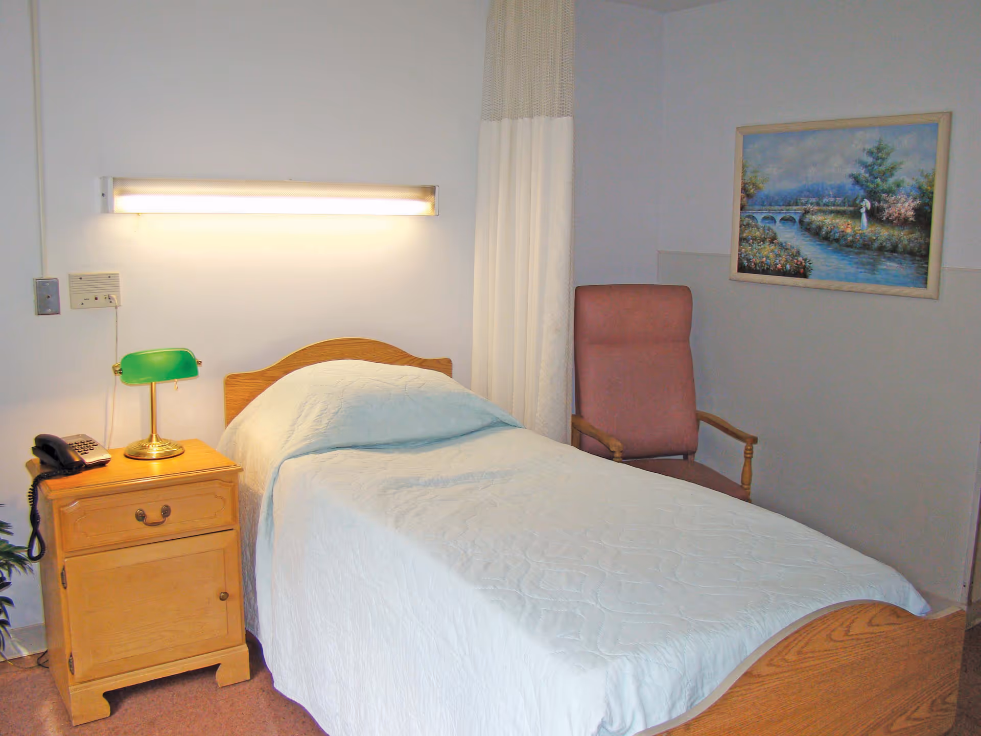 A small bedroom with a single wooden bed covered with a light blue quilt. Next to the bed is a wooden nightstand with a green lamp and a telephone. A pink upholstered chair is placed against the wall, and a painting of a river scene hangs above it. A white curtain is partially visible behind the chair.