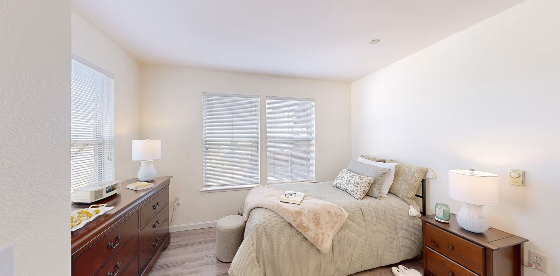 A bright bedroom with a neatly made bed featuring multiple pillows and a beige throw blanket. There are two wooden nightstands on either side of the bed, each with a white lamp. A wooden dresser with a lamp, a book, and a decorative tray is positioned against the left wall. The room has three windows with blinds, and the floor is a light wood laminate.
