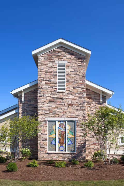 Exterior view of a building with a stone facade and a stained glass window depicting a religious figure. The building is surrounded by small trees and shrubs under a clear blue sky.