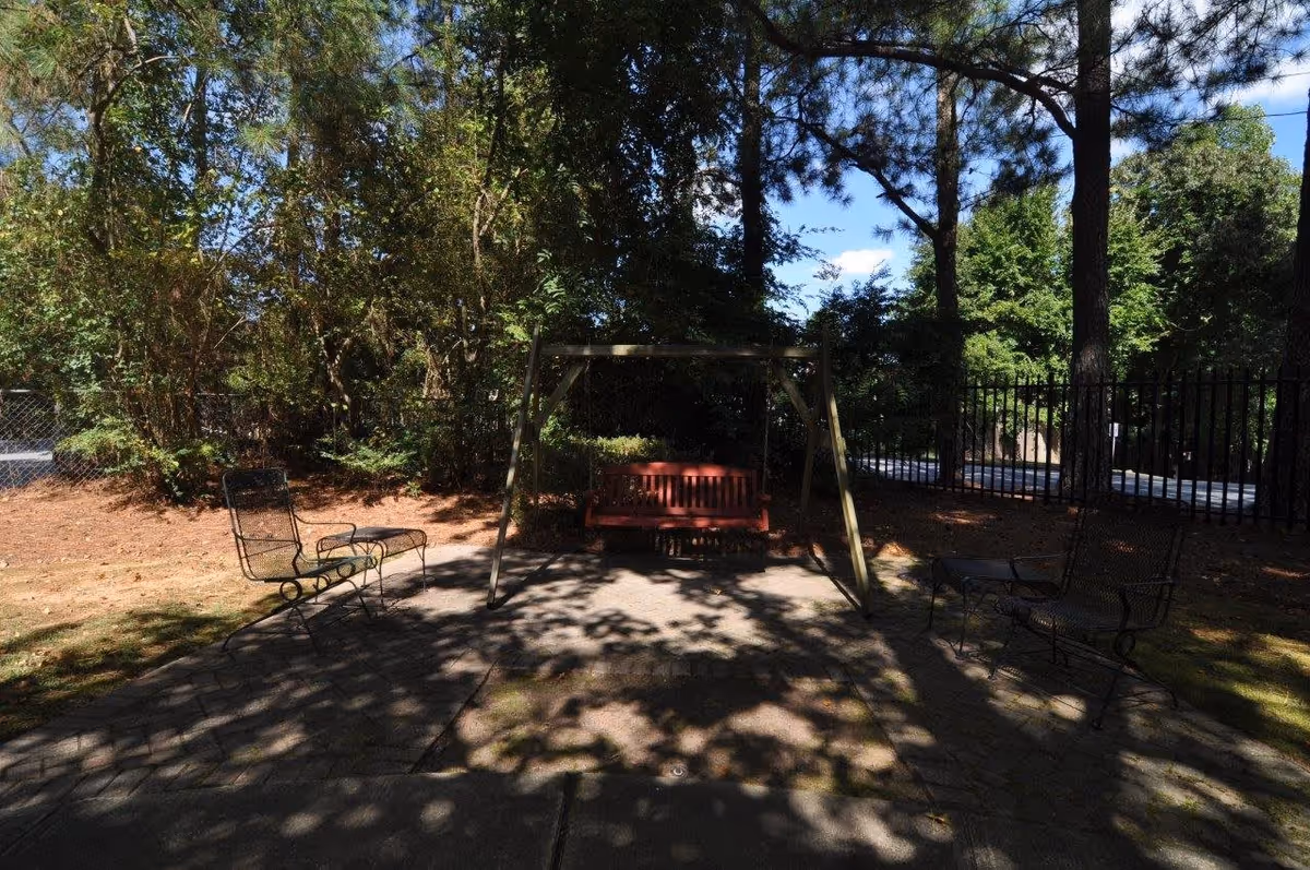 Outdoor seating area with a red wooden swing hanging from a wooden frame, surrounded by four black metal chairs on a paved surface. The area is shaded by tall trees and enclosed by a black metal fence, with dense greenery in the background.