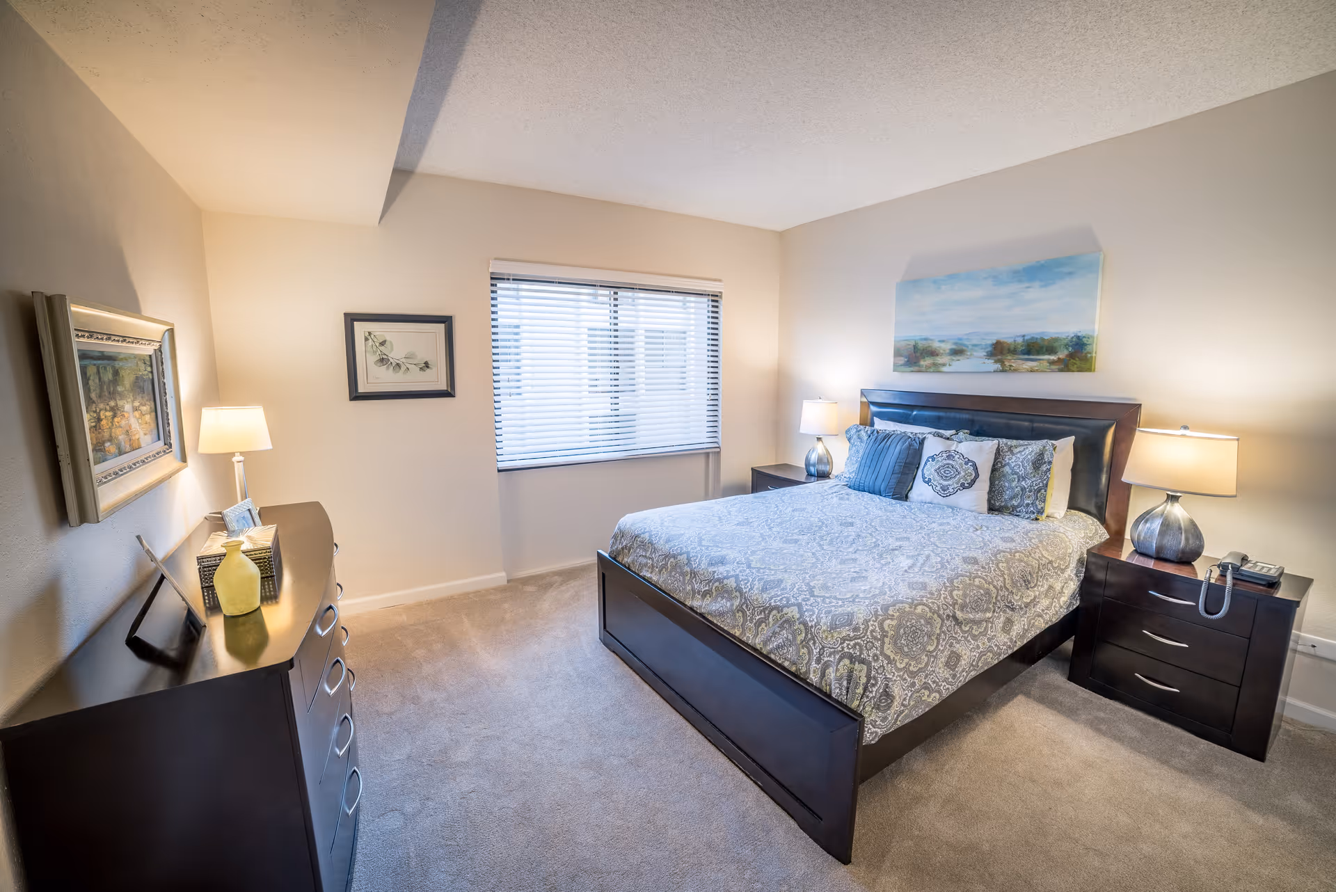 Light, furnished bedroom featuring a bed with patterned bedding, two nightstands with lamps, a dresser, and a window.