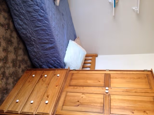 A small bedroom with a wooden bed frame, a blue quilted bedspread, a white pillow, and a wooden dresser with drawers. The room has beige walls and a carpeted floor with a floral pattern.