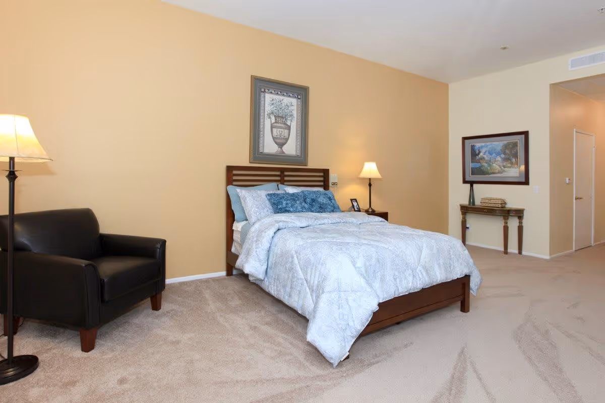 Spacious bedroom featuring a bed with blue bedding, a black armchair, two lamps, and framed artwork on beige walls.