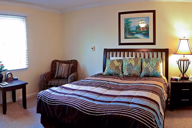 A cozy bedroom with a large bed featuring a striped comforter and leaf-patterned pillows. To the right of the bed is a nightstand with a decorative lamp and an alarm clock. On the left side of the room, there is a window with blinds, a small table with a clock and a flower vase, and a cushioned armchair with striped upholstery. A framed landscape painting hangs on the wall above the bed.