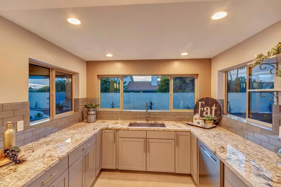 Modern kitchen with granite countertops, beige cabinets, a stainless steel sink under a large window, and decorative items including a wooden sign that says 'Eat', a small plant, and a bottle with grapes on the counter.