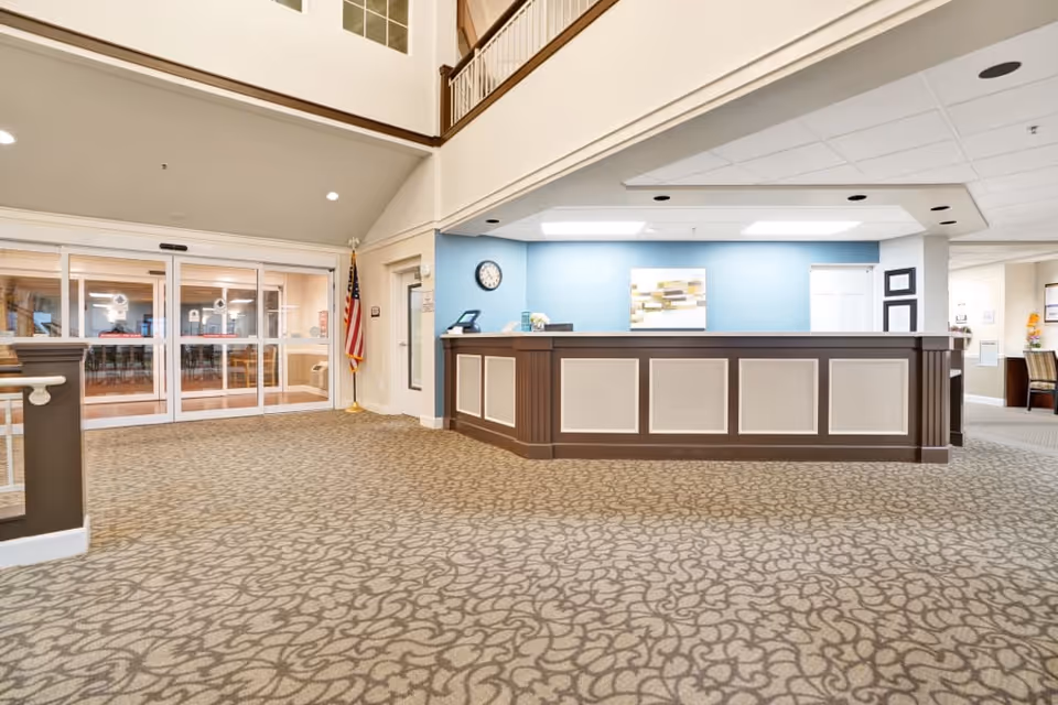 Reception area inside The Commons At Dallas Ranch facility with a large front desk, blue accent wall behind the desk, a clock, an American flag, and automatic sliding glass doors leading to another room.