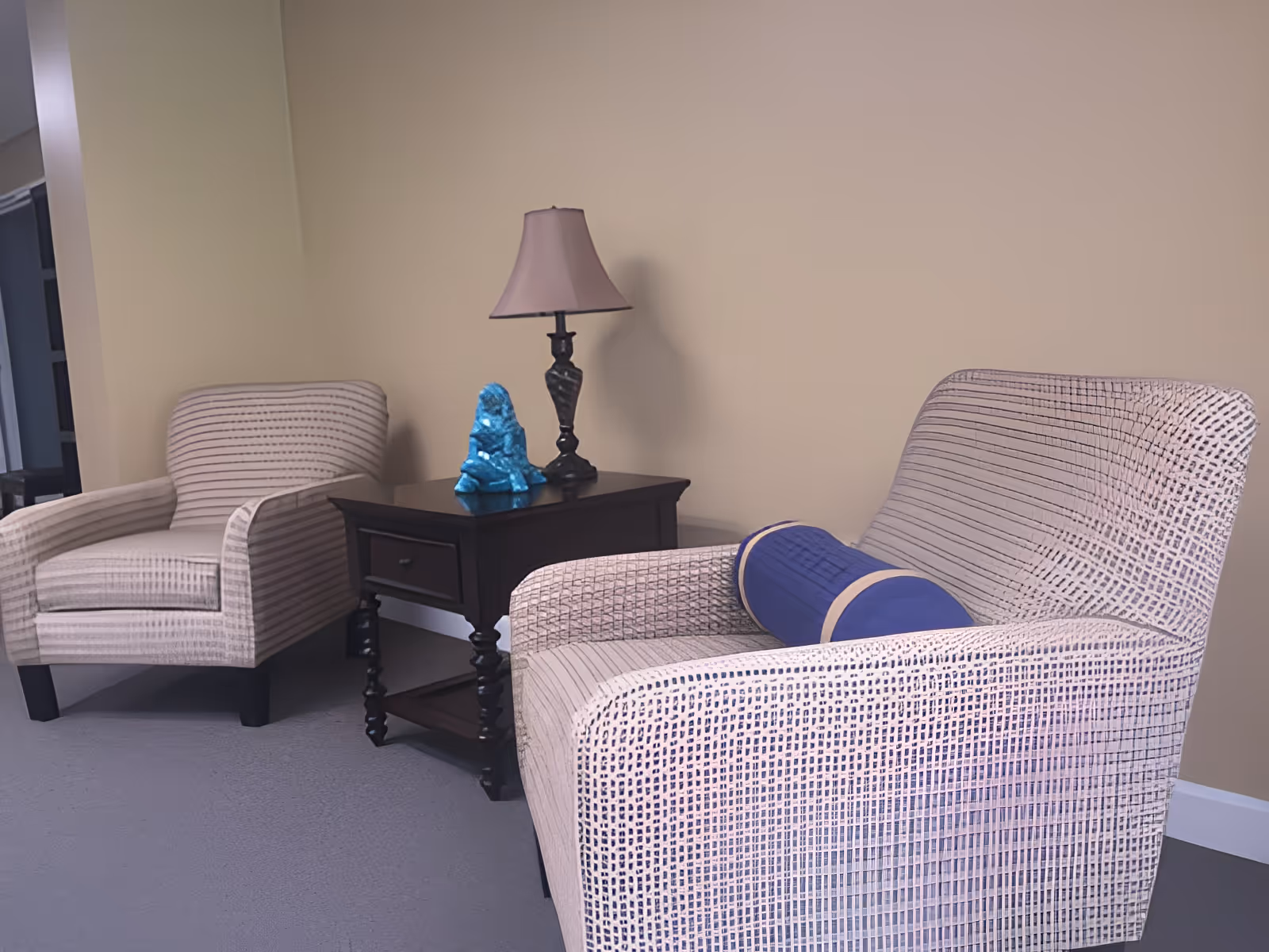 Two patterned armchairs with a blue cylindrical pillow on one, positioned around a dark wooden side table with a drawer. On the table, there is a decorative blue sculpture and a lamp with a beige shade. The setting is indoors with beige walls and carpeted floor.