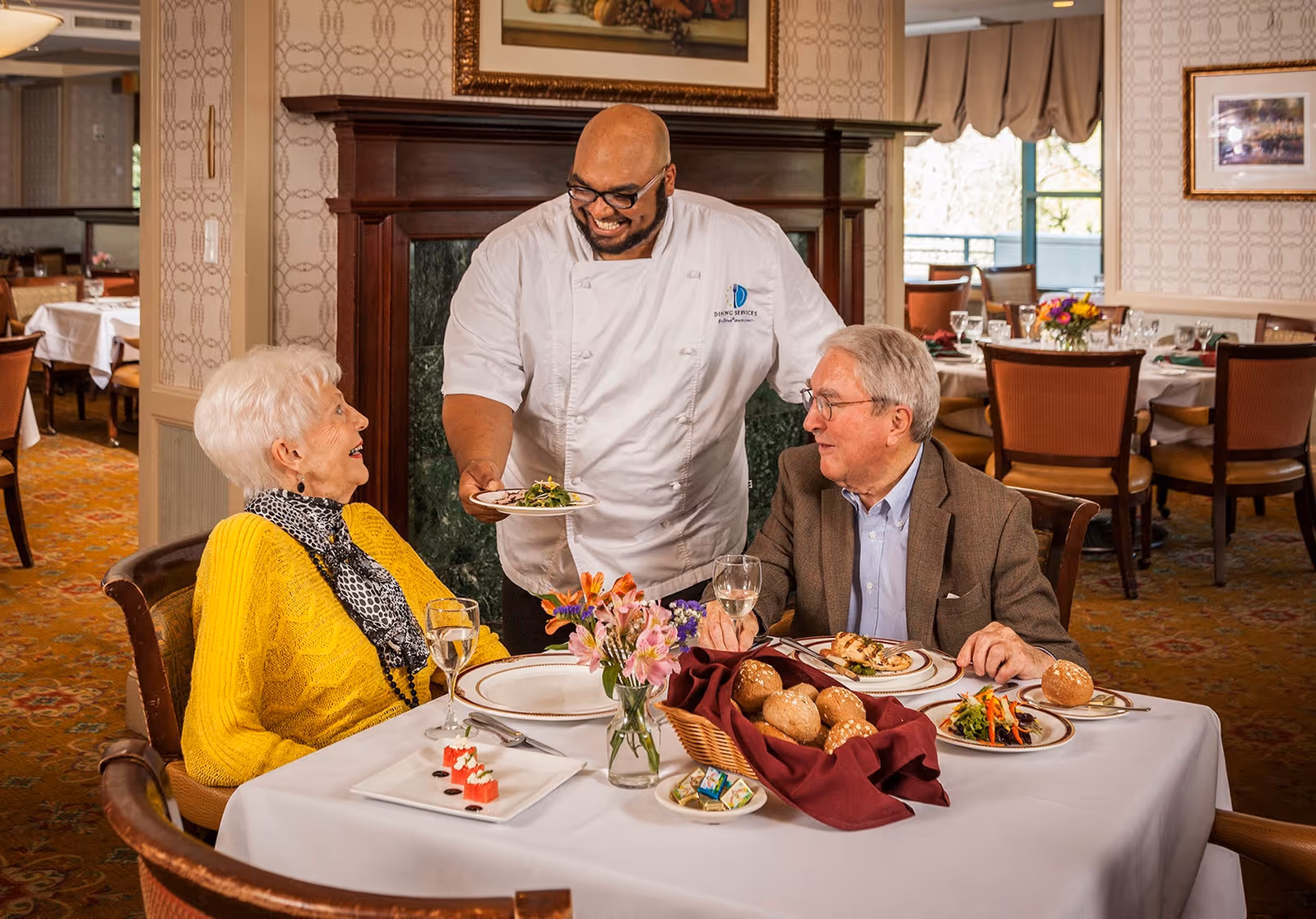 A chef serving a plate of food to an elderly couple seated at a dining table in a well-decorated restaurant. The table is set with plates, glasses, a basket of bread rolls, and a vase of flowers. The elderly woman is wearing a yellow sweater and a patterned scarf, while the elderly man is dressed in a brown blazer and glasses. The background shows more dining tables and chairs in a cozy dining room setting.