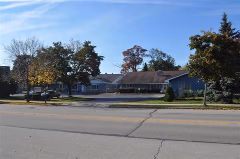 Exterior view of a single-story assisted living facility building with a parking area in front, surrounded by trees with autumn foliage under a clear blue sky.