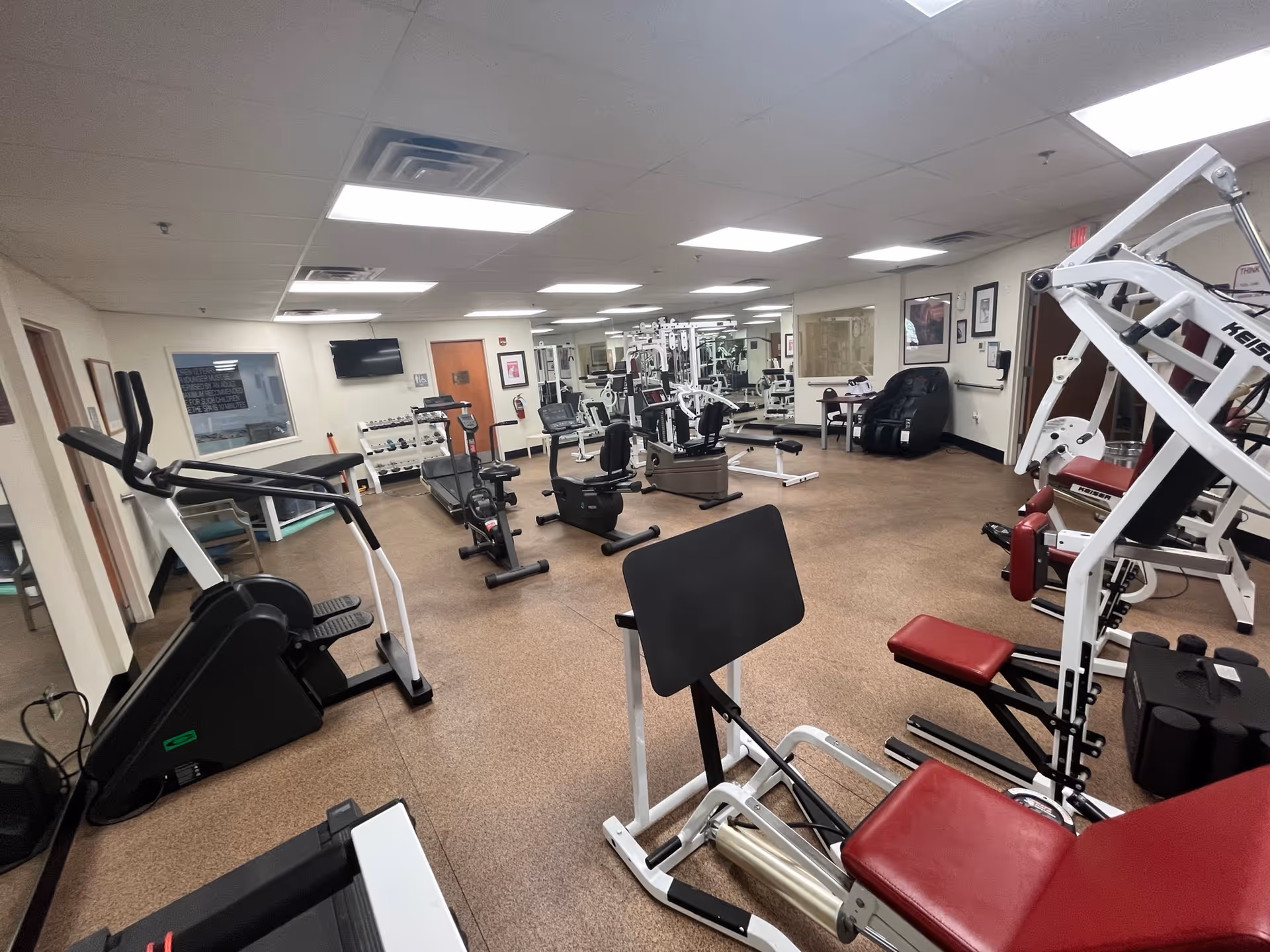 Interior view of a fitness room in a senior living facility with various exercise equipment including treadmills, stationary bikes, weight machines, and a massage chair. The room has a brown floor, white walls, and a ceiling with fluorescent lights. Mirrors cover one wall, and there is a television mounted on another wall.