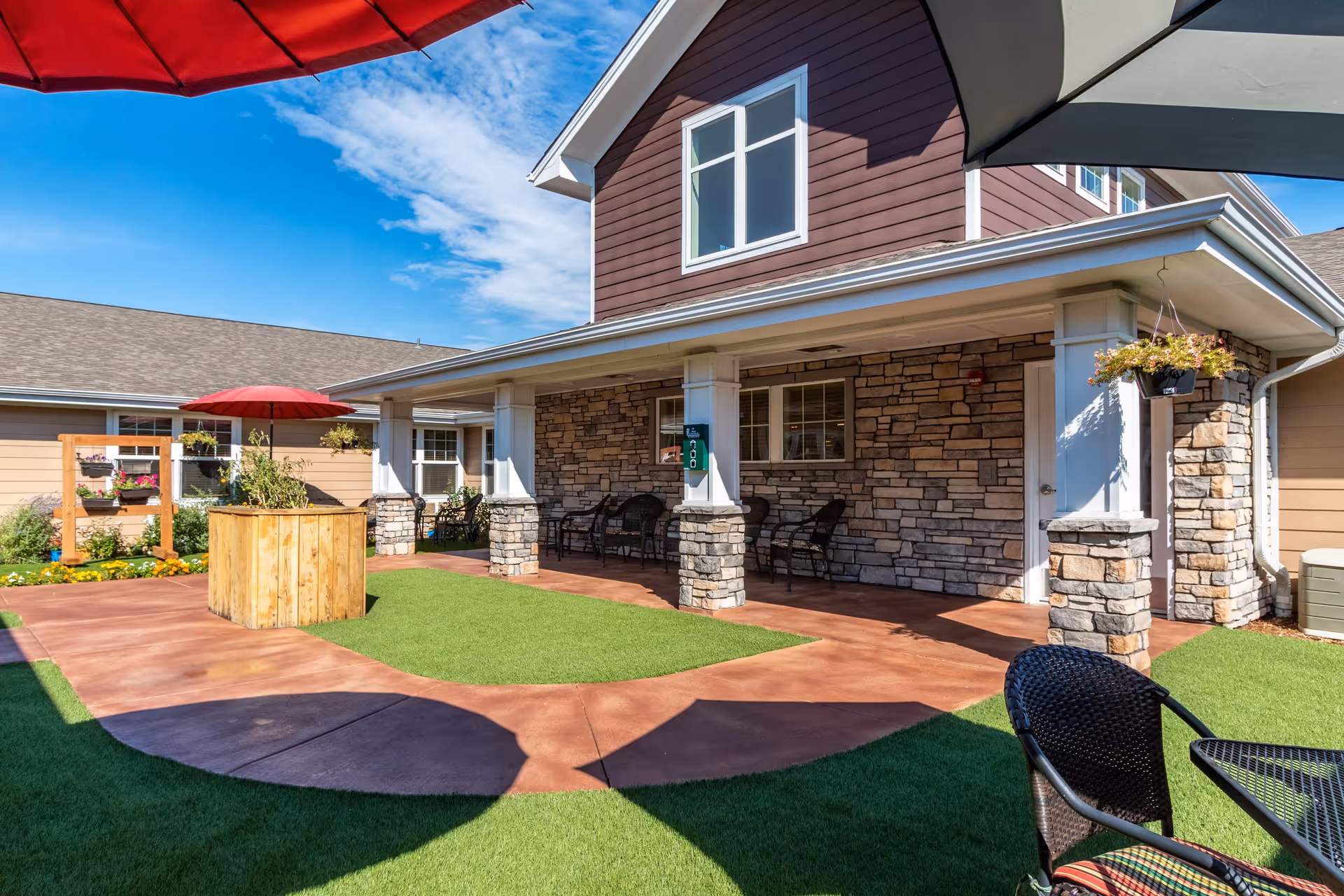 Outdoor courtyard area of a senior living facility with artificial grass, a concrete walkway, stone pillars supporting a covered porch, several chairs, hanging flower pots, and large red and gray umbrellas providing shade under a blue sky with some clouds.