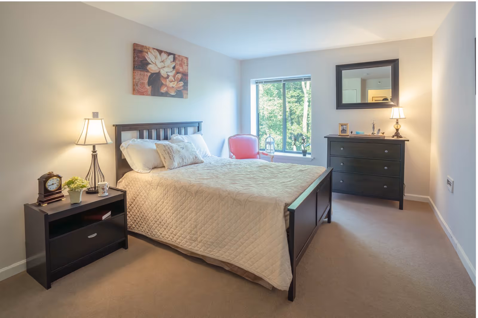 A well-lit bedroom with a large bed covered in a beige quilt, two pillows, and a decorative cushion. To the left of the bed is a black nightstand with a lamp, a clock, a small plant, and a cup. On the wall above the bed is a floral painting. To the right of the bed is a window with a view of green trees outside, a red armchair, and a black dresser with a mirror above it and a small lamp on top. The room has beige carpet and light-colored walls.