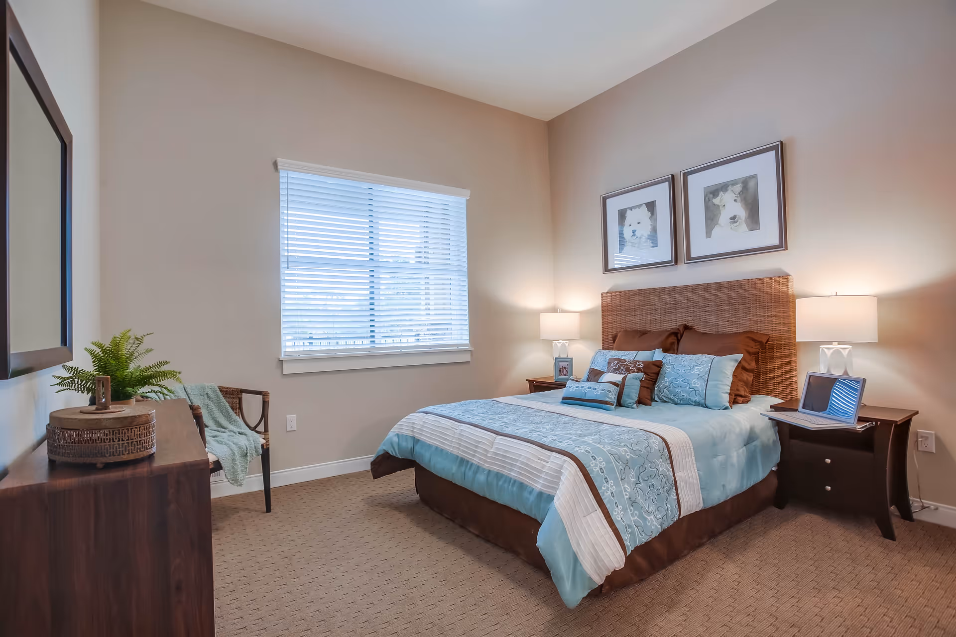 A neatly made bed with a blue and brown patterned comforter and multiple pillows in a bedroom. The bed has a woven headboard and is flanked by two dark wooden nightstands, each with a lamp. Above the bed are two framed pictures of dogs. A window with white blinds is on the left wall, and a dark wooden dresser with a plant and decorative items is on the left side of the room. The walls are painted beige and the floor is carpeted.