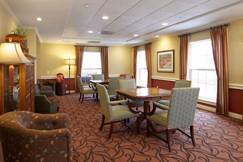 A well-lit senior living common area with multiple tables and cushioned chairs arranged for socializing or activities. The room features large windows with striped curtains, patterned carpet, a wooden cabinet with a lamp on top, and framed artwork on the walls.