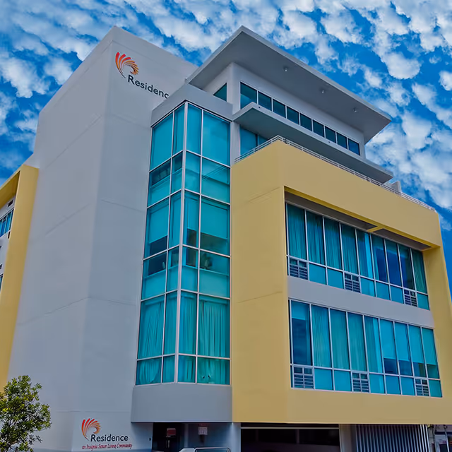 Exterior view of a modern multi-story senior living facility building with large blue-tinted windows and a yellow and white facade under a partly cloudy sky.