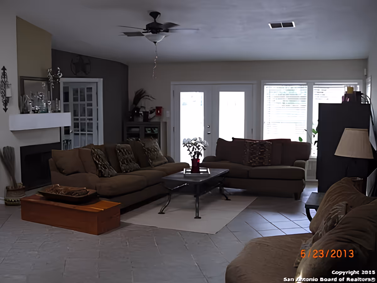A living room with two brown sofas arranged around a black coffee table on a light-colored rug. There is a fireplace on the left wall with decorative items on the mantel, a ceiling fan above, and large windows with blinds letting in natural light. A lamp and a cabinet are visible on the right side of the room.