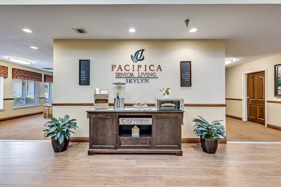 Interior view of a senior living facility reception area with a wooden cabinet holding a water dispenser, coffee supplies, and a microwave. The wall behind the cabinet displays the sign 'Pacifica Senior Living Skylyn'. Two potted plants flank the cabinet, and there are hallways visible on either side.