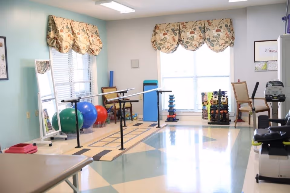Bright therapy/exercise room with parallel bars, exercise balls, hand weights, chairs, and large windows.