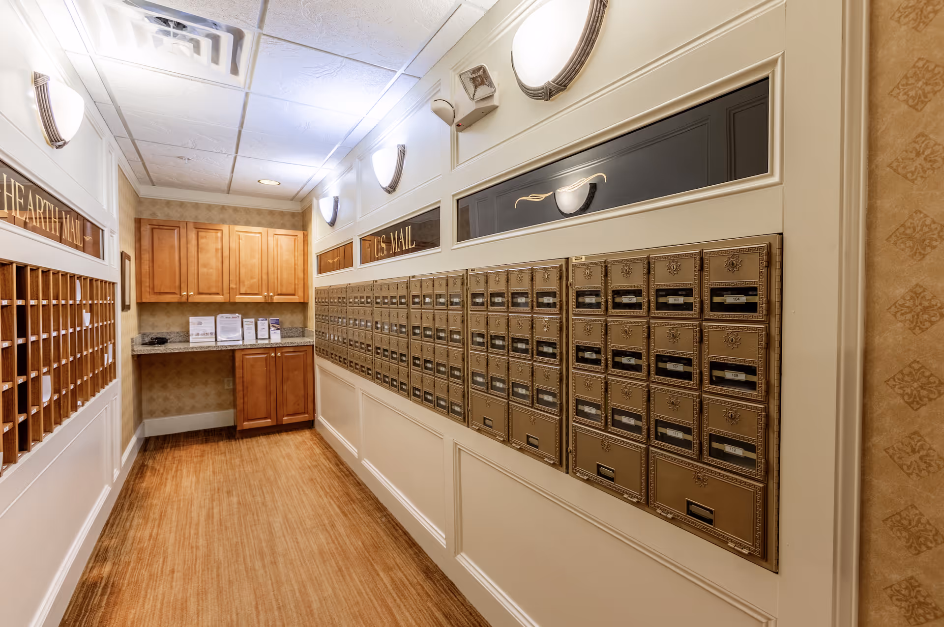 Interior view of a mailroom with multiple brass mailboxes mounted on the right wall and wooden cubbyholes on the left wall. There are wooden cabinets and a countertop at the end of the room, with soft lighting fixtures on the walls and a patterned wallpaper.