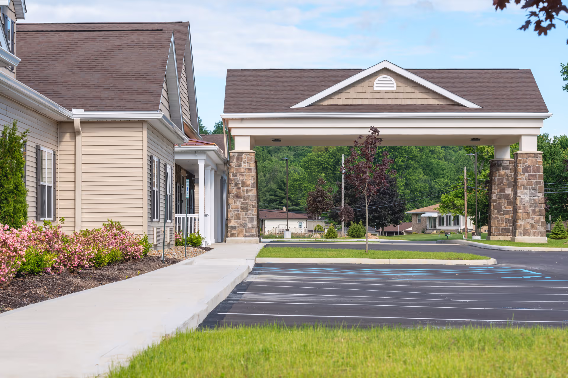 Exterior view of Tallmadge Health and Rehab facility showing a covered entrance with stone pillars, beige siding, a sidewalk, landscaped bushes with pink flowers, and an empty parking lot with handicap spaces.